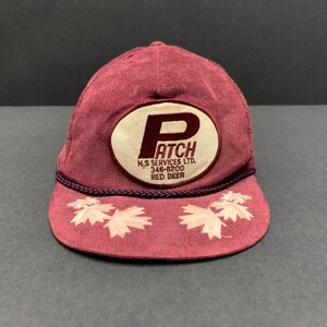 Pode incluir: Boné de beisebol de veludo cotelê bordô com um patch branco e bordô que diz "PATCH". A aba apresenta folhas de bordo bordadas. O boné tem um detalhe de corda bordô ao redor da aba.