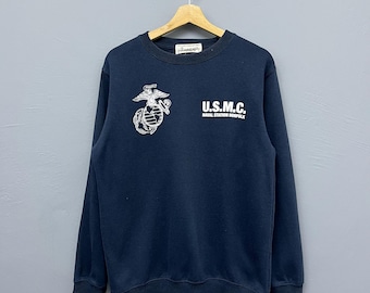 May include: Navy blue sweatshirt with the text "U.S.M.C. Naval Station Norfolk" in white. The sweatshirt also features a white emblem on the left chest. The sweatshirt is hanging on a wooden hanger.