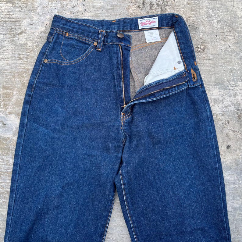 K&ouml;nnte beinhalten: Dunkelblaue Jeans aus Denim mit Rei&szlig;verschluss und Knopfverschluss. Die Jeans haben ein klassisches F&uuml;nf-Taschen-Design und eine gerade Beinpassform. Der Markenname "Wrangler" ist auf einem Etikett im Bund sichtbar.