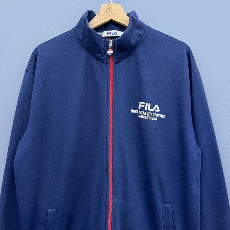 Puede incluir: Chaqueta deportiva azul marino con cremallera roja y el logo de Fila en el pecho y el cuello. La chaqueta tiene un cuello alto y dos bolsillos delanteros. El texto "FILA MODA NELLA VITA SPORTIVA NUMERO UNO" est&aacute; impreso en el pecho.