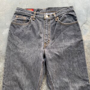 May include: Close-up of a pair of faded black denim jeans. The jeans have a button fly, classic five-pocket design, and a straight leg fit. The stitching is a contrasting tan color, and the jeans show signs of wear.