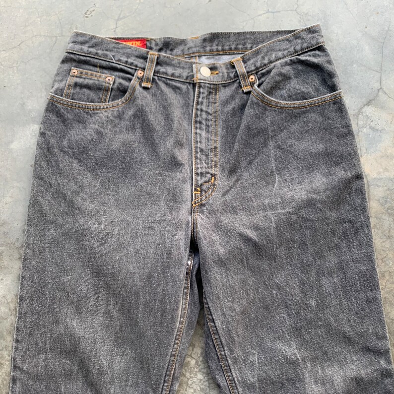 May include: Close-up of a pair of faded black denim jeans. The jeans have a button fly, classic five-pocket design, and a straight leg fit. The stitching is a contrasting tan color, and the jeans show signs of wear.