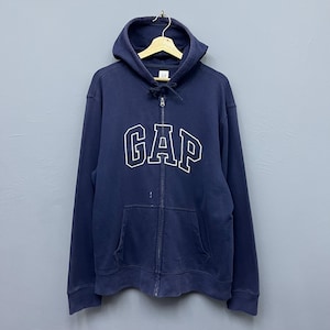 May include: A navy blue zip-up hoodie featuring the white outlined text "GAP". The hoodie has a drawstring hood, a front pocket, and a full-length zipper. The garment is hanging on a wooden hanger. The hoodie is a casual style.