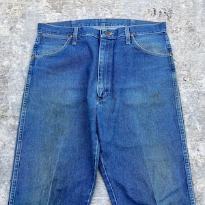 May include: Close-up of blue denim jeans. The jeans have a classic five-pocket design, with a button and zipper closure. The denim has a faded appearance, with some wear and tear visible. The jeans are a medium wash.