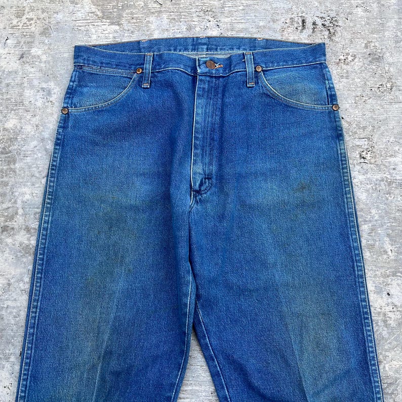 May include: Close-up of blue denim jeans. The jeans have a classic five-pocket design, with a button and zipper closure. The denim has a faded appearance, with some wear and tear visible. The jeans are a medium wash.