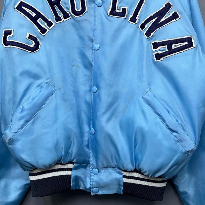 May include: Light blue satin jacket with the word "CAROLINA" in white lettering. The jacket has a snap-button closure, two front pockets, and a ribbed collar, cuffs, and hem with white stripes. A classic varsity style.