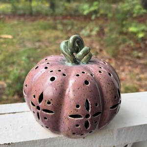 May include: A ceramic pumpkin sculpture in a dusty mauve color with dark speckles. The pumpkin features a glossy green stem and cut-out patterns including small circular holes and four-petaled flower shapes across its rounded surface.