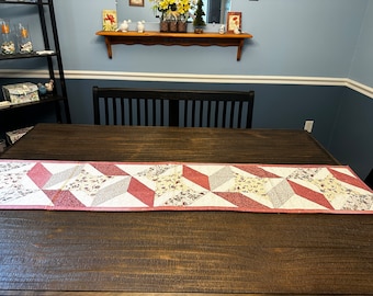 May include: A pink and white quilted table runner with a geometric pattern. The runner is on a dark wood table.