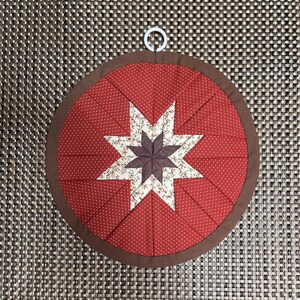 May include: A round, quilted hot pad with a red background and white polka dots. A star design is in the center, with a brown star in the middle and white floral print points. The edge is brown, and it has a white loop for hanging.