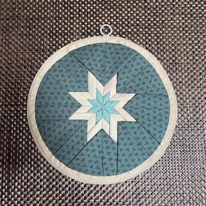 Könnte beinhalten: Ein runder, gesteppter Topfuntersetzer mit Sternmuster. Er hat einen teal-farbenen Hintergrund mit floralem Muster, einen zentralen Stern in Hellblau und Creme und einen cremefarbenen Rand. Ein Metallring ist zum Aufhängen angebracht.