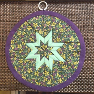 Könnte beinhalten: Ein runder, gesteppter Topflappen mit einem grünen Sternmuster in der Mitte. Der Stern ist von einem floralen Muster mit gelben und violetten Akzenten umgeben. Der Rand ist einfarbig lila, und es hat eine weiße Schlaufe zum Aufhängen. Durchmesser ca. 20 cm.