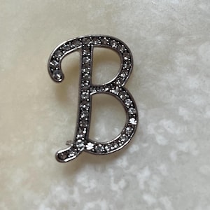 May include: A silver-toned letter "B" brooch, encrusted with small, clear gemstones. The brooch is in a cursive font and is set against a light-colored background. The gemstones sparkle, adding a touch of elegance.