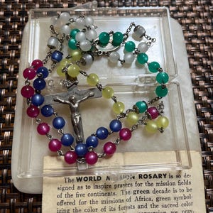 May include: A colourful rosary featuring beads in white, green, yellow, blue, and pink, presented in a clear plastic case. A silver crucifix is at the centre. A small card with text is visible below the rosary.