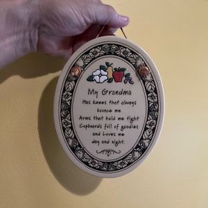 May include: Oval ceramic plaque with a floral border and the text "My Grandma." The plaque features a cream-coloured background with a decorative floral and fruit design at the top. A brown cord is attached for hanging.