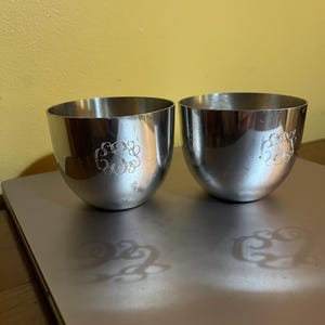 May include: Two silver cups with a monogram design. The cups are sitting on a silver surface.