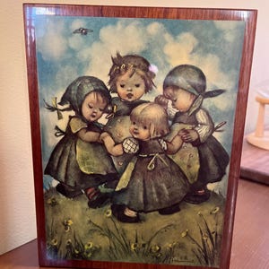 May include: A wooden music box with a colorful illustration of four children holding hands and dancing in a circle. The children are wearing traditional clothing and are surrounded by flowers and a blue sky. The music box is decorated with a brown wooden frame.