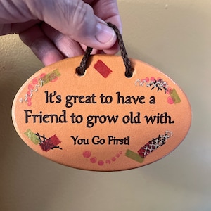 Może przedstawiać: Pomarańczowa, owalna tabliczka ceramiczna z tekstem "It's great to have a Friend to grow old with. You Go First!" w czarnym kolorze. Tabliczka ma brązowy sznurek do zawieszenia i jest ozdobiona kolorowymi szwami i farbą.