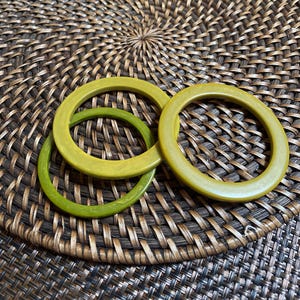 May include: Three green bangles in varying shades of green are arranged on a woven, brown surface. The bangles are circular and appear to be made of a solid material. The background is a textured, circular woven mat.