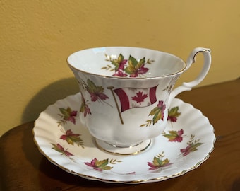 May include: A white teacup and saucer with a gold rim. The teacup has a red and white Canadian flag design with maple leaves. The saucer has a similar design with maple leaves.