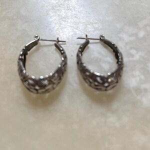 May include: A pair of silver-toned hoop earrings. Each earring has an oval shape with a detailed, openwork design. The earrings have a post and clasp closure. The background is a light, neutral color.