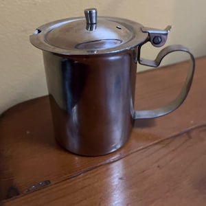 May include: A silver stainless steel creamer with a hinged lid and a handle. The creamer is sitting on a wooden surface.