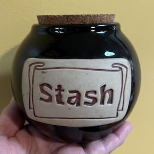 Op de afbeelding: Zwarte keramische pot met een kurken deksel en een beige label met de tekst "Stash" in bruin.