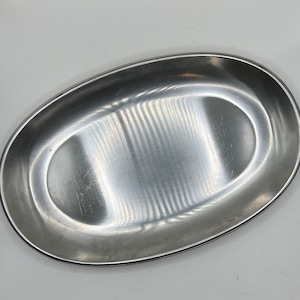 May include: A silver oval serving dish with a smooth, shiny surface. The dish has a slightly raised rim and a subtle, textured pattern.