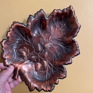 May include: A copper-colored leaf-shaped dish with a detailed design of veins and strawberries. The dish has a dark brown border and is held by a hand. The decorative dish is likely made of metal and is suitable for serving or display.