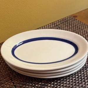 May include: A stack of four oval-shaped white plates with a dark blue border. The plates are stacked on top of each other, showcasing the clean design and color contrast. The plates are on a brown woven surface.
