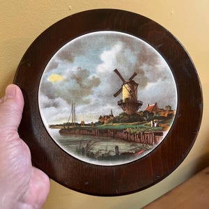 Könnte beinhalten: Runder dekorativer Teller mit einer gemalten Landschaftsszene mit einer Windmühle, Wasser und Gebäuden. Der Teller ist in einem dunkelbraunen Holzrahmen gefasst. Das Kunstwerk hat eine Vintage-Ästhetik.