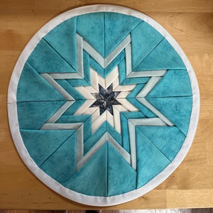 Könnte beinhalten: Ein runder, gesteppter Tischset mit einem Sternmuster. Der Stern besteht aus weißen, hellblauen und dunkelblauen Stoffen. Der äußere Ring ist helltürkis und der Rand ist mit weißem Stoff verziert.