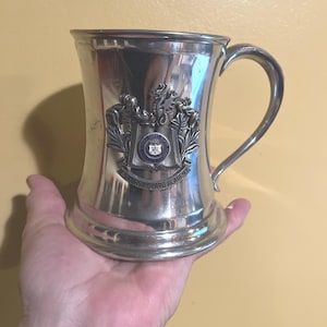 May include: A silver-colored metal mug with a curved handle. The mug features an embossed emblem with the text "U.S. COAST GUARD ACADEMY". The mug is held in a person's hand.
