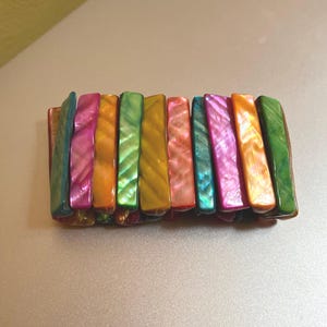 Könnte beinhalten: Ein farbenfrohes Armband aus rechteckigen, irisierenden Perlen. Die Perlen sind in den Farbtönen Grün, Rosa, Orange und Gelb gehalten. Das Armband besteht aus mehreren aneinandergereihten Perlen.