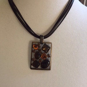 Può includere: Una collana con cordoncino in pelle marrone con un ciondolo quadrato con un design geometrico di pietre marroni e arancioni.