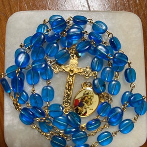 Könnte beinhalten: Eine blaue Glasrosenkranz mit goldenen Akzenten, mit einem goldenen Kreuz und einem kleinen ovalen Anhänger mit einem religiösen Bild. Der Rosenkranz liegt auf einer weißen Oberfläche.