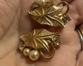 May include: A pair of gold-toned clip-on earrings. Each earring features a textured leaf design, faux pearls, and clear crystal accents. The earrings have a vintage aesthetic.