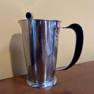 May include: A stainless steel pitcher with a black handle and a small lid. The pitcher has a cylindrical body and a rounded base. The handle is curved and attached to the side of the pitcher. The lid has a small, round knob.