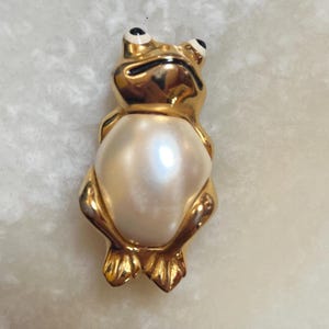 Peut inclure: Broche en forme de grenouille dorée avec un grand centre blanc nacré. La grenouille a des yeux noirs et blancs et des pieds dorés détaillés. Cette épingle unique est un accessoire fantaisiste.