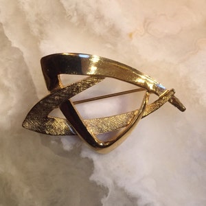 Könnte beinhalten: Eine goldfarbene Brosche mit geometrischem Design. Die Brosche ist aus Metall und hat eine strukturierte Oberfläche.