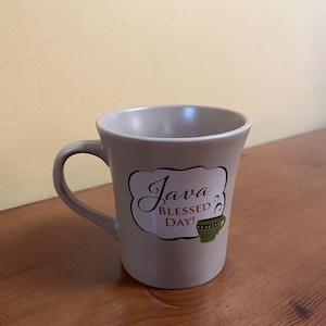 Puede incluir: Una taza de café de cerámica beige con un asa curvada. La taza presenta el texto "Java Blessed Day!" en una fuente decorativa dentro de un marco blanco y marrón, junto con una ilustración de taza de café verde.