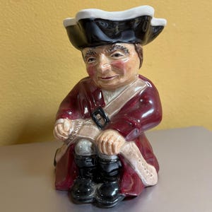 May include: A ceramic figurine of a man in a tricorn hat and burgundy coat. The figurine has a detailed face and is wearing black boots. The hat is black with a white trim. The figurine is a decorative item.