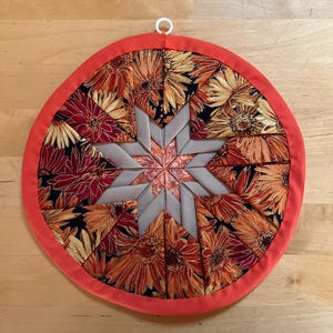 May include: A circular quilted potholder featuring a folded star pattern in the center. The design incorporates fabric with orange and yellow sunflower prints on a black background, solid beige fabric segments, and a bright orange fabric border.
