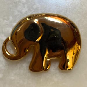 May include: A gold-colored elephant figurine with a smooth, reflective surface. The elephant is depicted in a stylized, rounded form, with visible details like the trunk and ears. The figurine is likely a decorative item.
