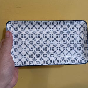 Peut inclure: Plat rectangulaire en céramique avec un fond blanc et un motif floral bleu répétitif. Le plat a une bordure bleue et est tenu par une main. Ce plat décoratif convient pour servir des amuse-gueules ou de petits plats.