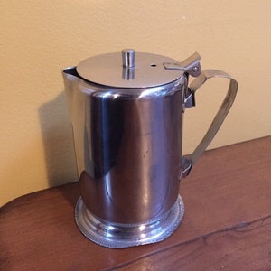 May include: A stainless steel pitcher with a lid and handle. The pitcher has a cylindrical body and a decorative base. The lid is round with a small handle. The handle is attached to the side of the pitcher.