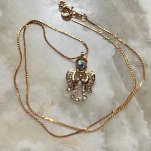 Pode incluir: Um colar de corrente de ouro com um pingente de anjo de cristal. O anjo tem uma pedra azul no centro.
