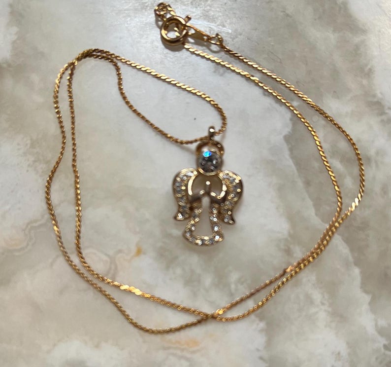 Pode incluir: Um colar de corrente de ouro com um pingente de anjo de cristal. O anjo tem uma pedra azul no centro.