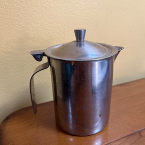 Könnte beinhalten: Ein silberner Edelstahl-Milchkännchen mit einem Klappdeckel und einem Griff. Der Milchkännchen steht auf einer Holzoberfläche.