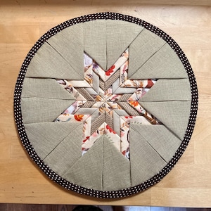 Può includere: Tovaglietta rotonda trapuntata con un motivo a stella. La stella centrale presenta motivi floreali e geometrici nei toni del beige, marrone e arancione. Le sezioni circostanti sono beige tinta unita e il bordo è rifinito con un tessuto a pois marrone.