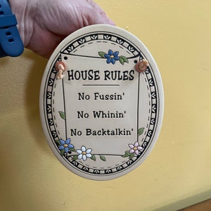 May include: A white oval ceramic sign with a blue and white floral border. The sign reads "House Rules" and lists three rules: "No Fussin', No Whinin', No Backtalkin'".